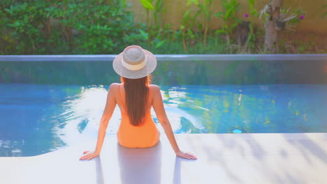 Back-view-of-a-female-tourist-sitting-near-the-swimming-pool,-wearing-orange-swimwear-and-a-sun-hat-,-soaking-her-feet-in-the-pool-water-enjoying-her-summer-holidays