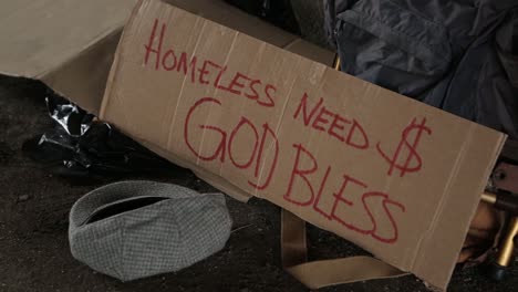 Nahaufnahme-Eines-Obdachlosenschilds-Aus-Pappe,-Das-Um-Geld-Bittet,-Während-Ein-Barmherziger-Samariter-Geld-In-Den-Hut-Steckt