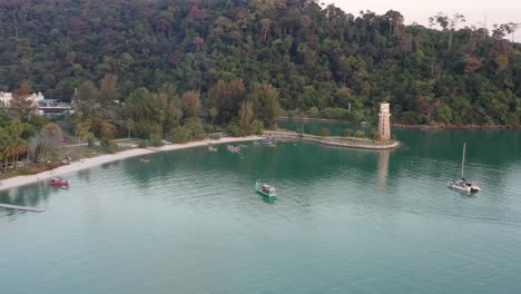 Rückwärtsflug-über-Der-Straße-Von-Malakka-Mit-Blick-Auf-Den-Leuchtturm-Perdana-Quay,-Der-Auf-Einem-Vorgebirge-Mit-Wunderschöner-Naturlandschaft-Und-Auf-Dem-Wasser-Festgemachten-Yachten-Und-Fischerbooten-Liegt