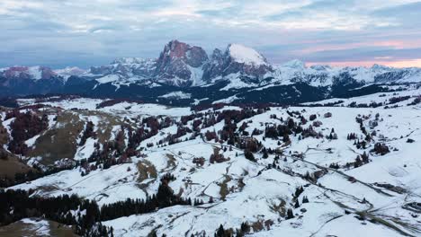 Backwards-aerial-shot-of-snowy-winter-landscape-in-front-of-Langkofel-and-Plattkofel-mountains-at-alpine-meadow-Seiser-Alm---Alpe-di-Siusi-in-the-Dolomites,-South-Tyrol,-Italy-at-sunset