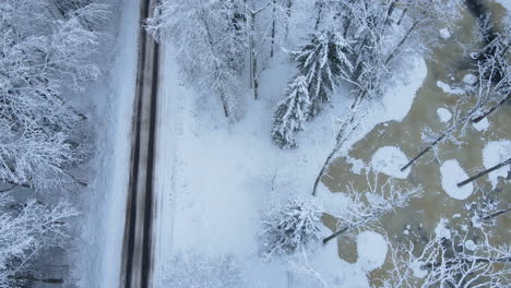 Aerial-view-of-an-empty-snowy-road-in-snow-covered-forest,-on-a-cloudy,-winter-day---drone-flying-back-along-the-road,-top-down-view