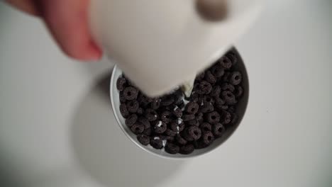 Milk-is-poured-into-the-bowl-with-chocolate-cereal,-drops-of-milk-falling-to-the-cocoa-cereal-breakfast,-top-down-view