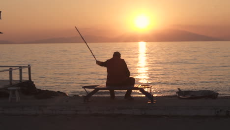 Anciano-En-El-Paseo-Marítimo-Sentado-En-Un-Banco-Con-Una-Ruta-De-Pesca-A-Cámara-Lenta