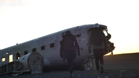 Travelers-walking-towards-crashed-and-abandoned-airplane-on-black-sand-beach-in-sunset