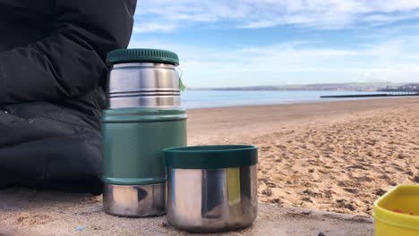 Drinking-hot,-steaming-tea-from-a-flask,-on-a-beach