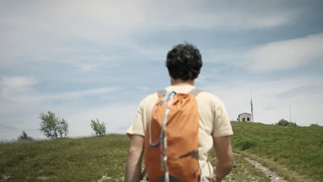 Hiker-walking-up-a-hill-towards-the-radio-tower-on-mountain-Slavnik,-camera-tracking-from-behind,-clouds-on-the-blue-sky