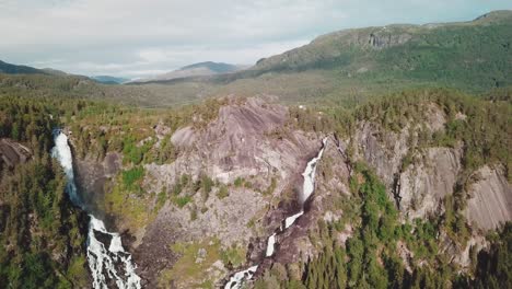 Backwards-drone-shot-over-two-large-waterfalls-and-a-forest-in-the-background-in-Norway,-shot-in-4K