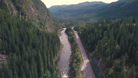 Crane-down-tilting-up,-high-angle-drone-shot-over-a-river-next-to-a-road-in-a-forest