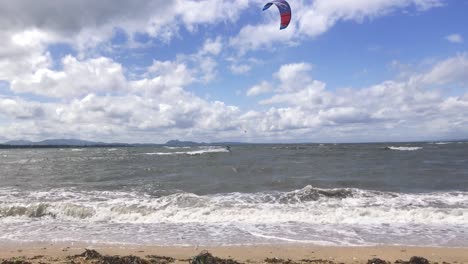 A-fixed-shot-of-waves-breaking-on-a-sandy-beach-with-kite-surfers-moving-through-the-shot-in-the-background,-on-a-windy-summer-day-|-Portobello-beach,-Edinburgh-|-Shot-in-HD-at-cinematic-24-fps
