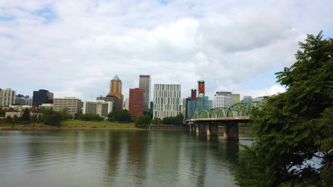 4K-Trucking-right-to-left-reveal-from-behind-a-tree-to-Hawthorne-Bridge-crossing-Willamette-River-toward-downtown-Portland,-Oregon-skyline-with-mostly-cloudy-sky-then-obscured-by-another-tree