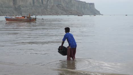 Junger-Pakistanischer-Junge,-Der-Plastikbehälter-In-Richtung-Meer-Trägt,-Um-Sich-Am-Strand-Von-Gwadar-Zu-Füllen
