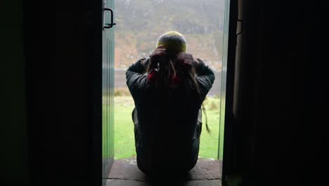 A-man-with-dreadlocks-and-wearing-hiking-gear-walks-into-shot-and-sits-on-a-step-in-a-narrow-doorway-of-a-bothy-in-the-Highlands-of-Scotland-to-enjoy-the-view-outdoors