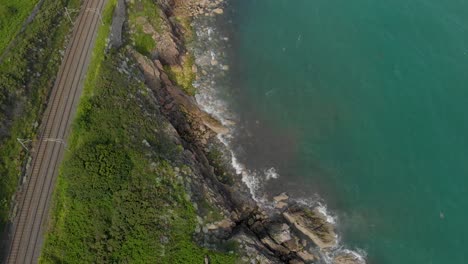 Top-down-drone-view-of-Irish-rail-road-near-the-green-sea-on-a-bright-sunny-day-while-waves-hit-the-shore