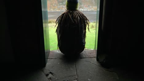 A-man-with-dreadlocks-and-wearing-hiking-gear-walks-into-shot-to-sit-on-a-step-in-a-narrow-doorway-of-a-bothy-in-the-Highlands-of-Scotland-to-enjoy-the-view-outdoors
