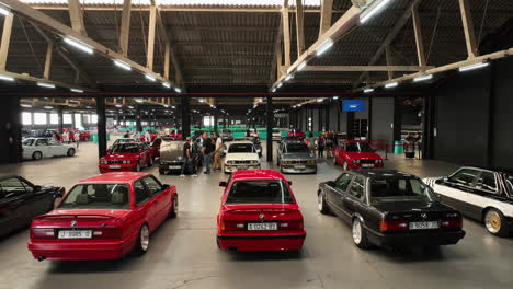 Colourful-retro-BMW-e30-vehicles-parked-at-warehouse-showcase-meeting,-aerial-view-dolly-right