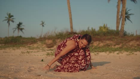 Schöne-Indische-Frau,-Die-Ein-Rotes-Sommerkleid-Trägt-Und-Mit-Ihren-Händen-Auf-Den-Sand-Zeichnet,-An-Einem-Tropischen-Strand,-Mit-Palmen-Im-Hintergrund