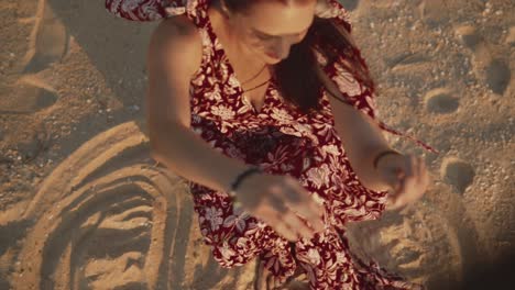 Beautiful-Indian-woman-wearing-a-red-summer-dress,-moving-sand-with-her-hands-and-letting-it-blow-in-the-wind,-on-a-tropical-beach