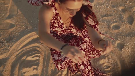 Bella-Mujer-India-Con-Un-Vestido-Rojo-De-Verano,-Moviendo-Arena-Con-Las-Manos-Y-Dejándola-Soplar-En-El-Viento,-En-Una-Playa-Tropical