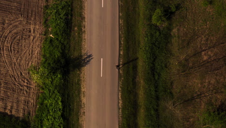 Aerial-drone-view-of-a-woman-running-on-the-side-of-a-countryside-empty-road,-on-a-sunny-day