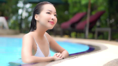Young-asian-tourist-standing-at-the-edge-of-a-swimming-pool-with-wet-her,-looking-around-and-smiling