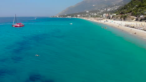 Anchored-boats-on-turquoise-sea-water-near-long-beach-on-beautiful-coastline-in-Dhermi,-Albania