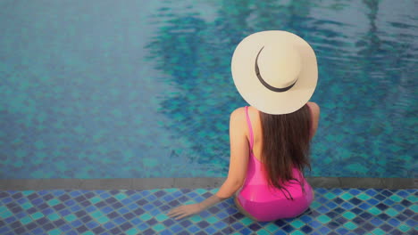 With-her-back-to-the-camera,-a-young-woman-in-a-pink-one-piece-bathing-suit-and-sun-hat-sits-on-the-first-step-of-a-hotel-swimming-pool