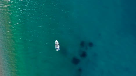 Boat-floating-on-clear-turquoise-sea-water-near-pristine-beach-where-people-sunbathe-in-Mediterranean