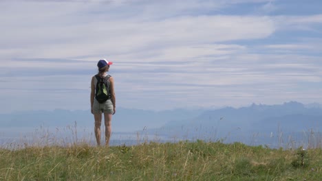 Una-Excursionista-Solitaria-Se-Encuentra-En-La-Cima-De-Una-Montaña-Mirando-La-Vista-De-Un-Gran-Lago-Alpino-Y-Los-Picos-Alpinos-Circundantes