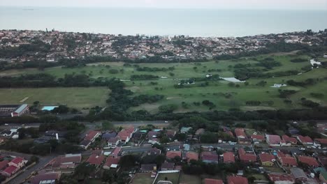 Aerial-footage-from-a-drone-of-residential-homes-with-a-road-with-moving-traffic-overlooking-a-water-tower-shopping-centre-and-a-golf-course-with-a-green-grassy-park