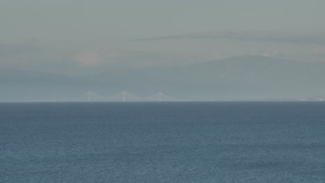 Long-range-telephoto-panning-shot-of-a-faraway-bridge-with-mountains-in-the-background-and-sea-in-the-foreground