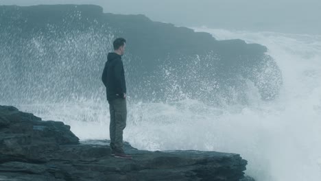 Man-standing-at-the-cliff-with-waves-crashing-in-the-background