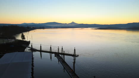 Drone-flying-above-a-stunning-lake-and-dock,-perfect-scenery-during-a-sunrise,-surrounded-by-mountains-in-the-distance