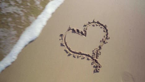 Heart-shape-drawing-in-the-sand-at-the-beach-is-getting-washed-away-by-a-sea-wave,-view-from-above