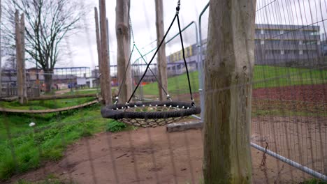 Empty-children-playground-closed-and-deserted-during-the-Covid-19-Coronavirus-pandemic-lockdown-in-the-UK,-big-swing-swaying-slowly-in-the-wind,-surrounded-by-metal-fences,-on-a-cloudy-day