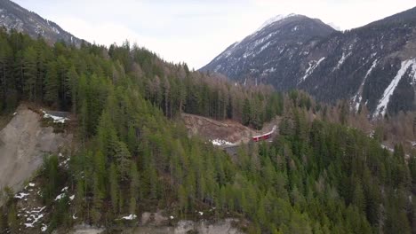 Aerial-view-of-the-famous-Bernina-train-moving-on-a-railroad-in-between-trees