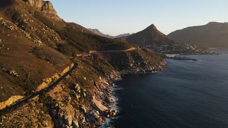 Drone-shot-of-coastal-road-with-cars-driving-on-it-next-to-cliff-leading-down-to-a-rocky-coastline-during-sunset
