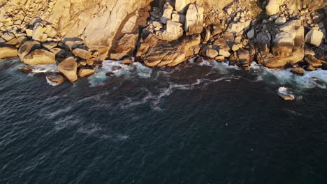 Cinematic-drone-shot-of-rocky-coastline-with-waves-crashing-against-the-boulders-and-seagulls-flying