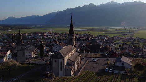 Aerial-orbit-shot-of-a-church-illuminated-by-the-sunlight-in-the-countryside-of-Vionnaz,-Switzerland