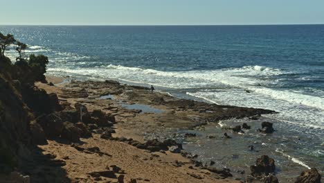 Lonely-person-walking-on-rocky-coastline-with-sea-waves-in-Sardinia,-distant-view