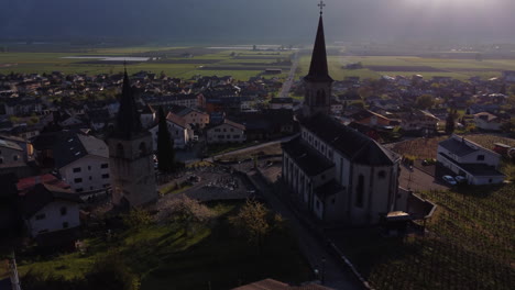 Slow-aerial-tilt-up-shot-next-to-a-church-in-Vionnaz,-Switzerland-with-sun-rays-in-frame