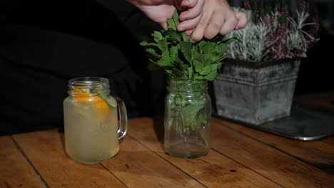 Hand-Des-Barkeepers,-Der-Frische-Grüne-Minze-In-Alkoholischen-Cocktail-Auf-Holztisch-Hinzufügt