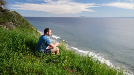 A-young-man-traveler-sitting-down-and-enjoying-nature-in-a-grassy-field-on-the-edge-of-a-beach-cliff-overlooking-the-ocean-view-of-Santa-Barbara,-California-SLOW-MOTION