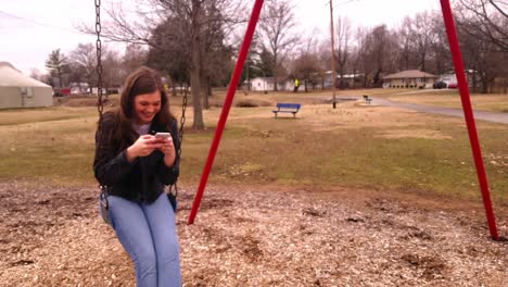 4k-footage-of-a-beautiful,-young,-brunette-college-teenager-smiling-and-laughing-while-checking-message-using-her-smart-phone-while-sitting-in-a-swing-set-at-a-city-park