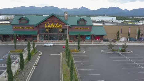 Aerial-flyover-of-a-Cabela's-store-and-parking-lot-in-Anchorage,-Alaska-showing-many-recreational-vehicles-parked-with-cars-moving,-people-walking,-and-mountains-in-the-background