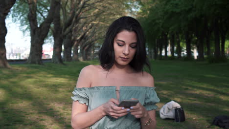 Confident-and-attractive-young-brunette-girl-in-a-casual-top,-uses-her-smartphone,-smiles-happily-to-the-text-messages,-looks-around-while-walking-in-a-park-with-a-line-of-trees-as-background