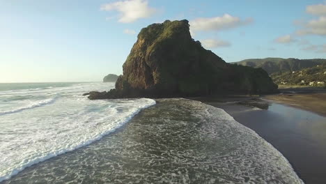 Aerial-tracking-shot-to-the-toward-the-heroic-Lion-Rock-as-white-water-washes-upon-the-sand-at-Piha-Beach,-Auckland-New-Zealand