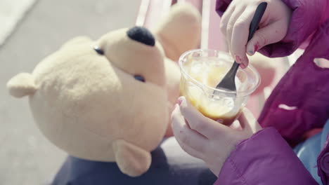 Close-up-of-little-girl-with-blue-jumper-nail-polish-and-teddy-bear-beside-her-holding-a-sundae-ice-cream-sitting-outdoor-4k