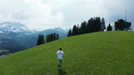 smooth-drone-shot-of-guy-walking-and-running-on-a-green-grass-hill-with-stunning-mountain-scenery,-entlebuch-switzerland