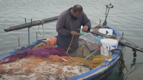 Hábil-Pescador-Griego-Reparando-Redes-En-Un-Pequeño-Barco-De-Pesca-Tradicional-De-Madera