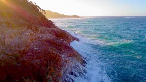 Aerial-view-of-a-rocky-cliff-coastline-and-turquoise-blue-water-waves-splashing-at-sunset